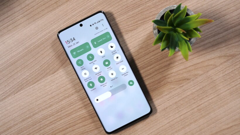 Элементы быстрого управления на смартфоне OnePlus 12OnePlus, лежащем на деревянной поверхности рядом с растением.