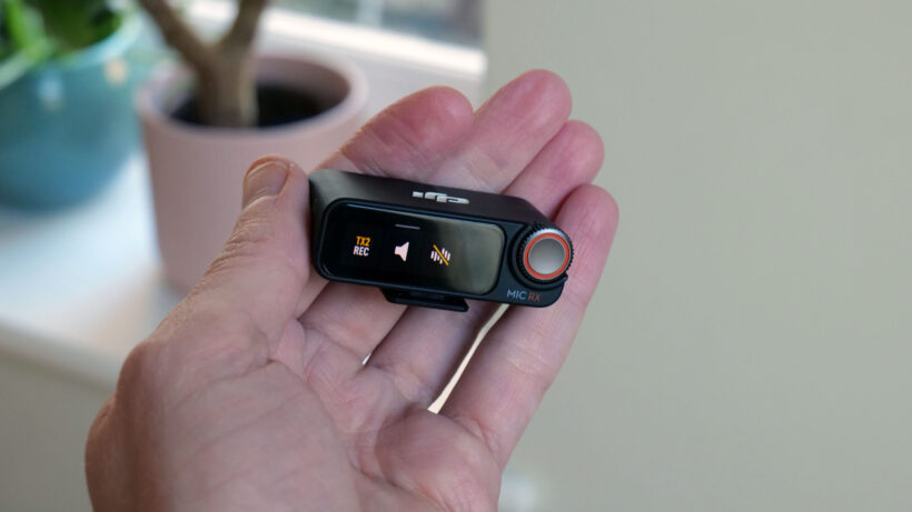DJI Mic 5 см: в руке держится беспроводной передатчик DJI Mic 2 с включенным дисплеем.
